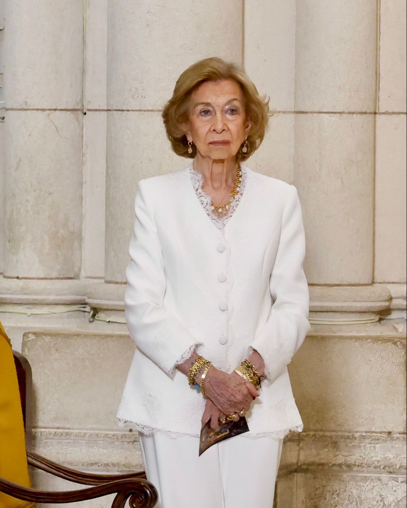 La Reina Sofía de España, con traje blanco y joyas, sostiene un objeto.