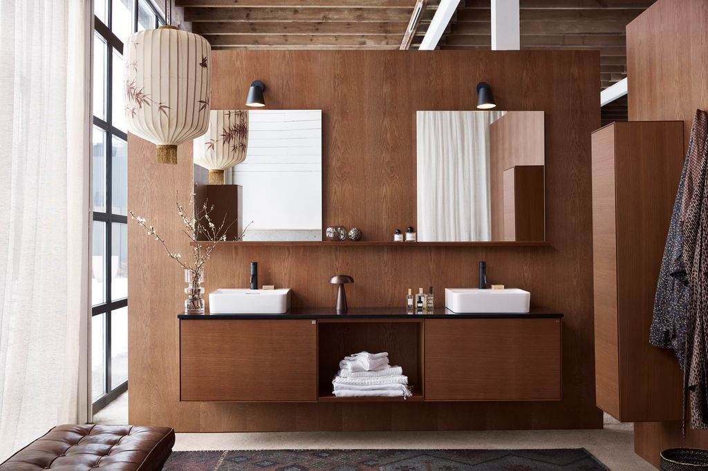 Baño de estilo japonés con paredes de madera oscura, muebles de madera, butaca de cuero marrón, lámpara de papel de estilo japonés, espejos de baño, dos lavabos, alfombra, cortinas, mueble alto en madera, albornoz colgando de una percha, jarrón de cristal 