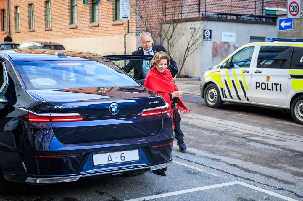 Queen Sonja de Noruega, con una bufanda roja, sale de un auto BMW, acompañada por un hombre en traje.