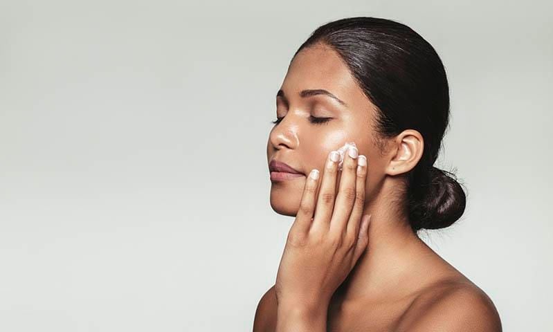 Hermosa mujer joven con los ojos cerrados mientras se aplica la crema hidratante para la cara limpia.