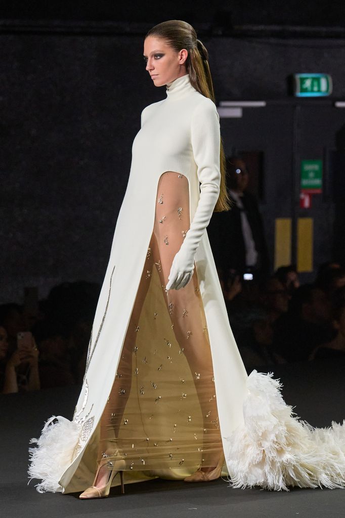 Modelo en pasarela con vestido blanco y dorado, tacones altos y guantes blancos.