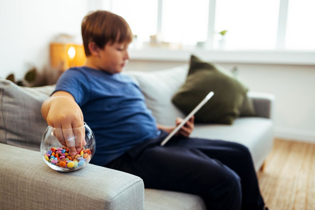 Niño con sobrepeso con la tablet en el sofá y tomando chocolate