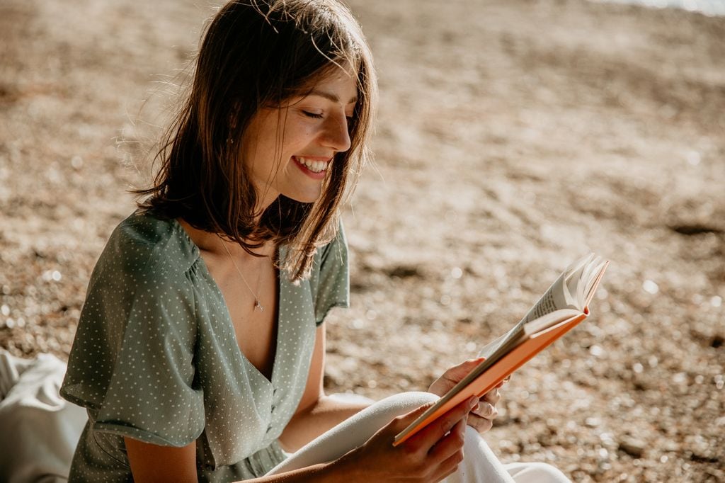 Mujer joven con vestido vaporoso en verde leyendo un libro naranja sobre la arena, muy sonriente
