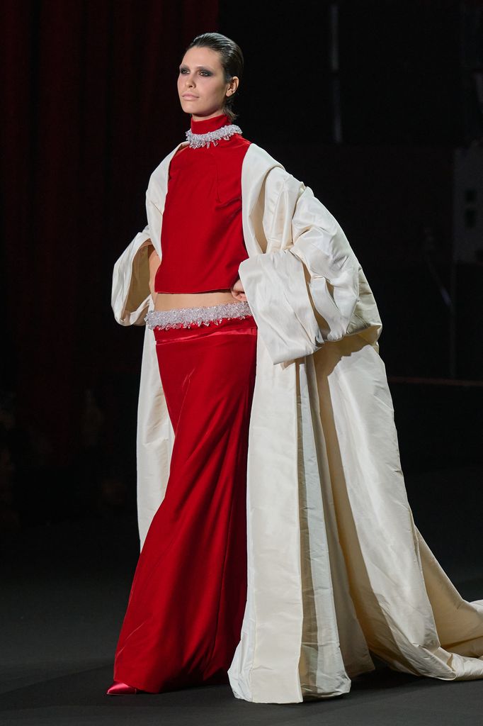 Modelo en pasarela con traje rojo y capa blanca en evento de moda.