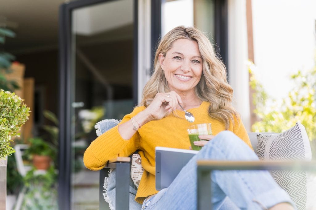 "Retrato de una mujer madura feliz sentada en la terraza con una tableta digital mientras disfruta de un batido verde."