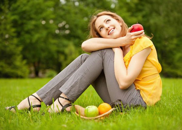 Mujer sentada en césped con cesta de frutas, sosteniendo una manzana.