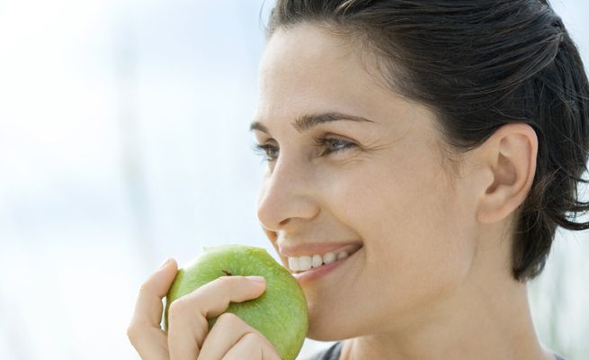 Mujer con cabello corto comiendo una manzana verde, sonriendo.