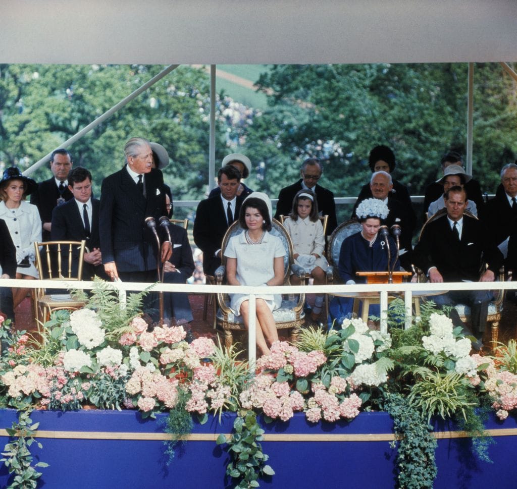 Ceremonia de inauguración del monumento a John F. Kennedy en el Reino Unido en 1965: Jacqueline Kennedy está sentada en primera fila con la reina Isabel II y el duque de Edimburgo. Justo detrás de Jackie se puede ver al Robert F. Kennedy, que también sería asesinado, y a la izquierda de la imagen se puede ver al Joan Kennedy junta su marido, Ted.