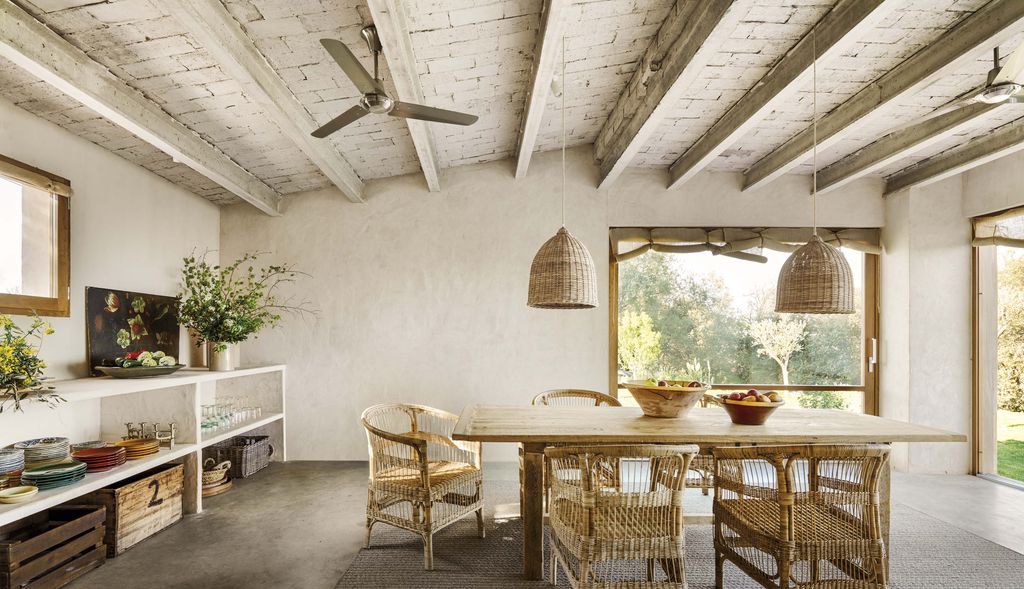 Comedor de estilo rústico, con vigas de madera pintadas de blanco, mesa de madera, sillas y lámparas de techo de fibras naturales, ventilador de techo, cajas de madera, jarrón con flores y cuadro