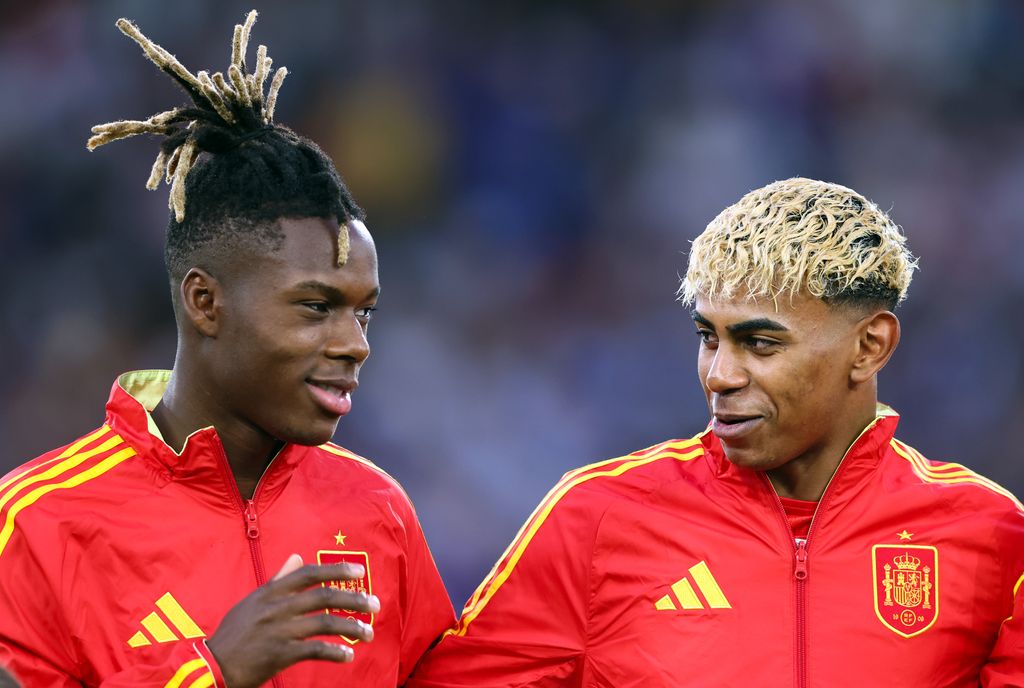 Dos jugadores de fútbol con chaquetas rojas y amarillas conversan en el campo.