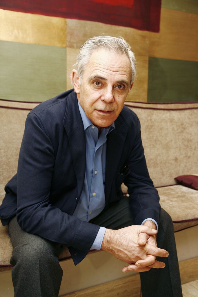 Hombre mayor con cabello blanco, traje azul y camisa celeste, sentado en sofá.
