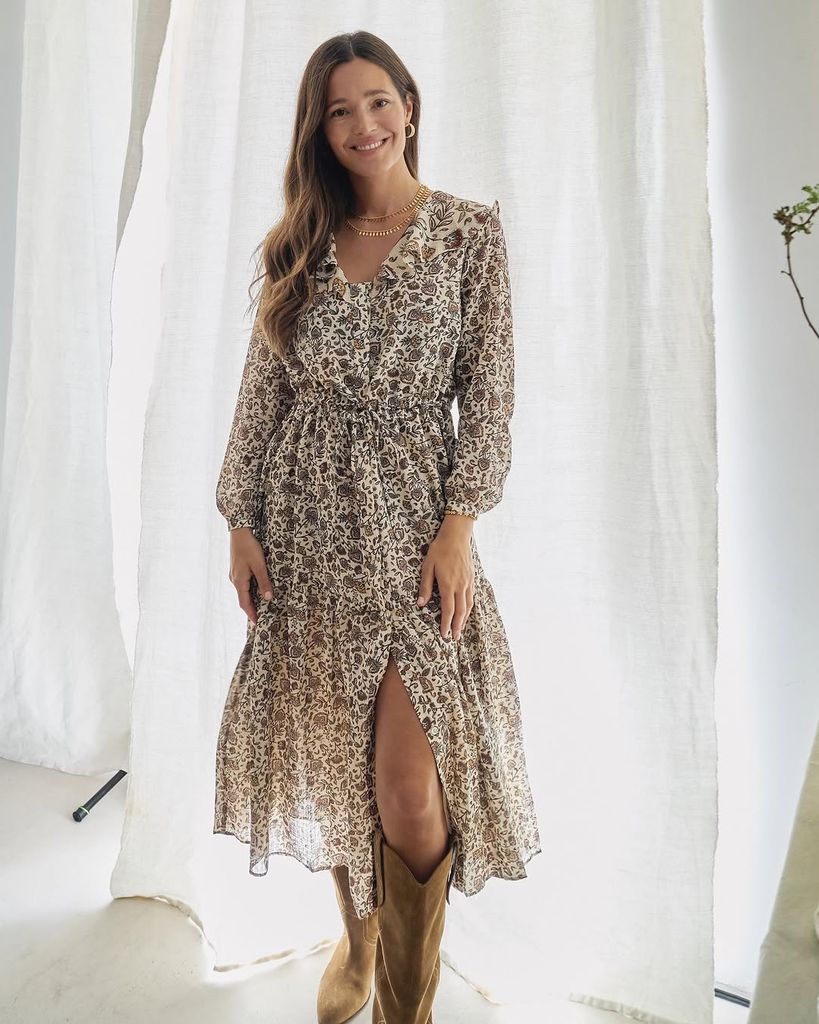 Mujer con cabello largo, sonriendo, vestida con un vestido floral y botas.