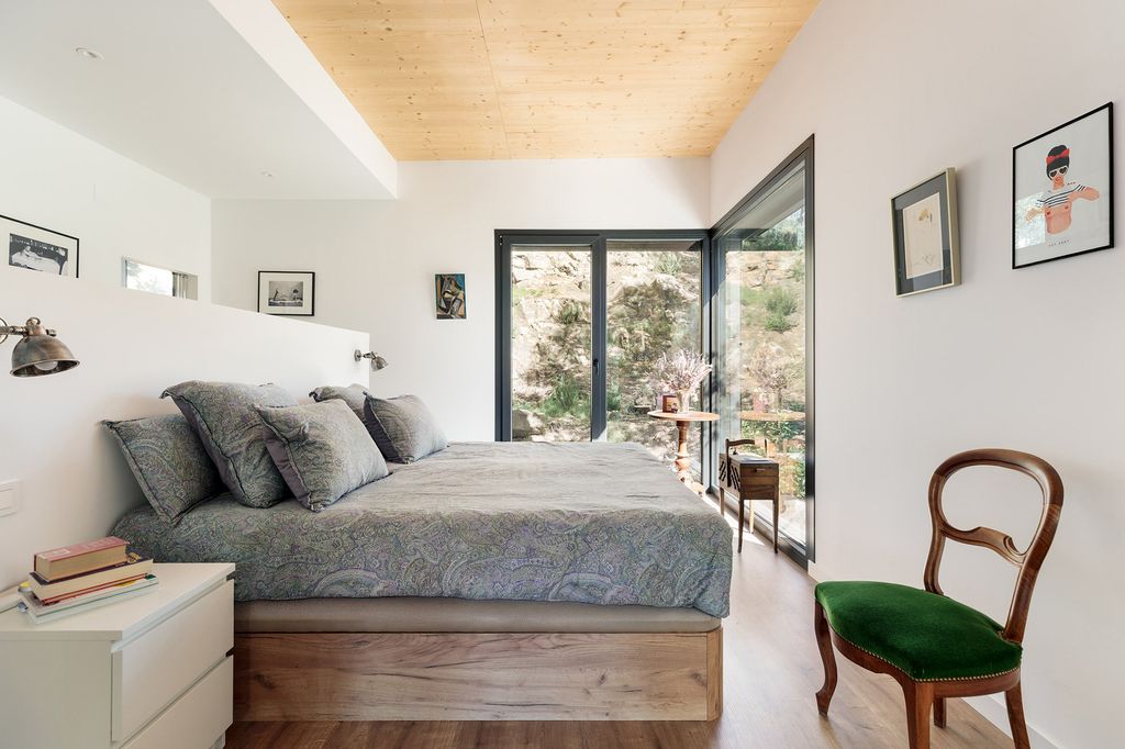 Dormitorio con cabecero de obra, mesita de noche blanca, suelo de madera, sillas antigua con el asiento de terciopelo verde, salida a terraza, techo de madera 