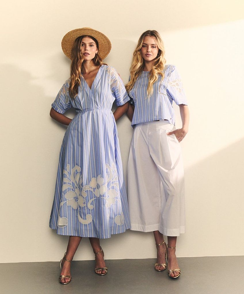 Dos mujeres posan juntas, una en vestido azul y otra en blanco.
