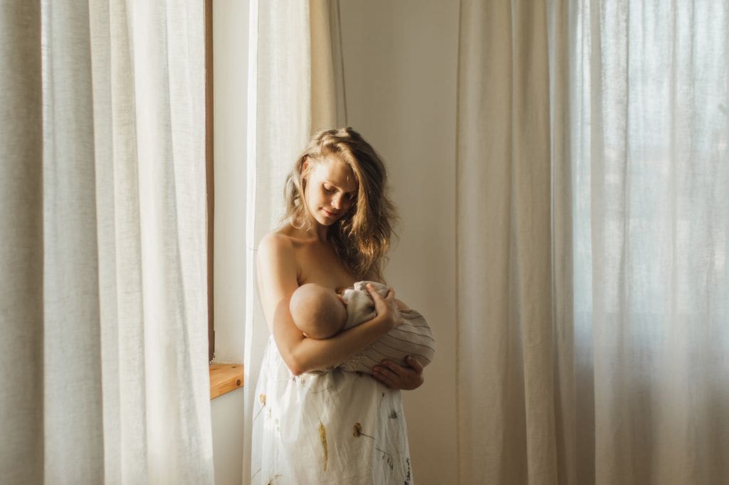 Mujer da el pecho a su bebé junto a la ventana