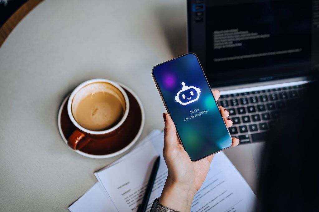 Mujer con un móvil en la mano, usando una web o app de inteligencia artificial, con un café y el portátil sobre la mesa