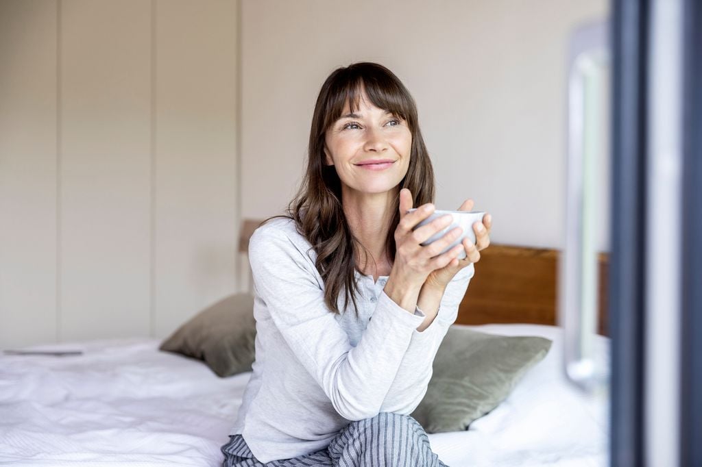 Mujer morena sonriente sentada en su cama con una taza de café