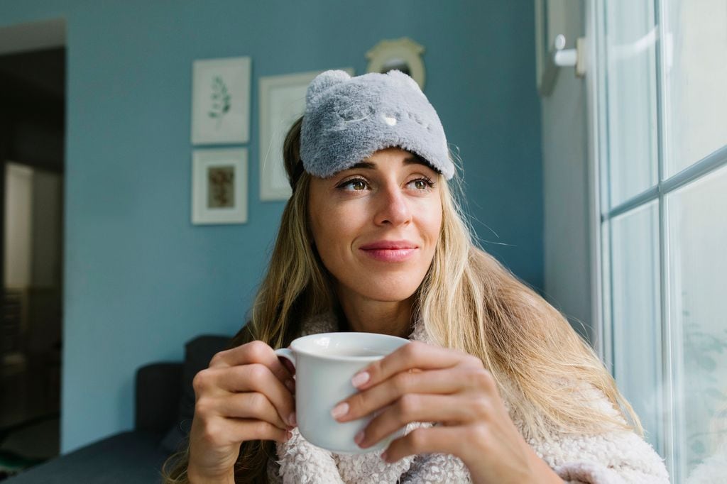 Mujer con antifaz y una taza de té mirando por la ventana