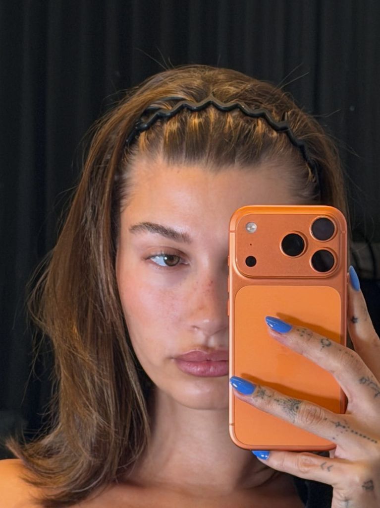 Hailey Bieber con diadema en zigzag, teléfono con carcasa naranja y manicura azul