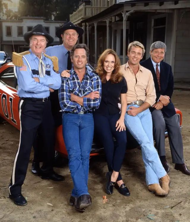 En la foto, de izquierda a derecha: James Best (como Rosco P. Coltrane), Rick Hurst (como el ayudante Cletus Hogg), Tom Wopat (como Luke Duke), Catherine Bach (como Daisy Duke), John Schneider (como Bo Duke), Ben Jones (como Cooter Davenport).