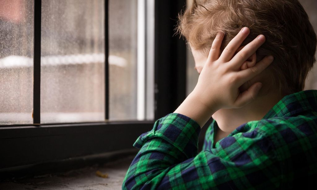 Little boy standing behind the window in sad mood. Sad Teenager looking in the Window and closing his ears with hands. Unhappy child in a plaid shirt. Alone at home. Upset.