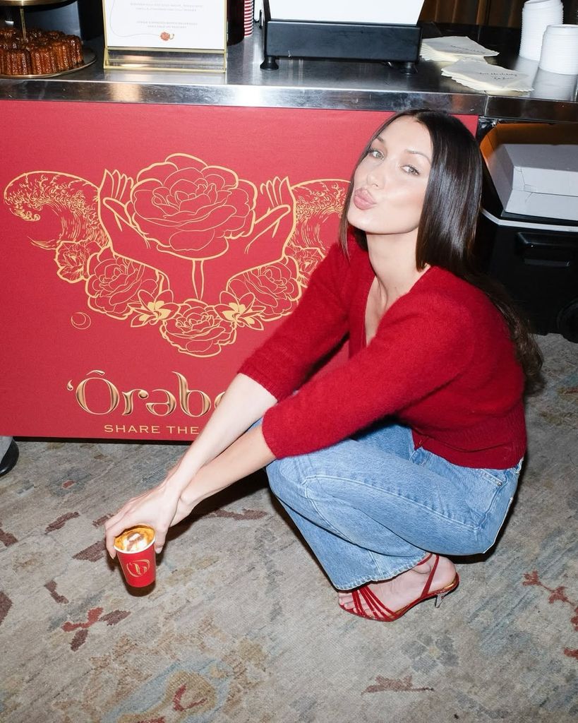 Bella Hadid agachada con un jersy rojo y jeans, posando con gesto de beso frente a un mostrador decorado con ilustraciones florales.