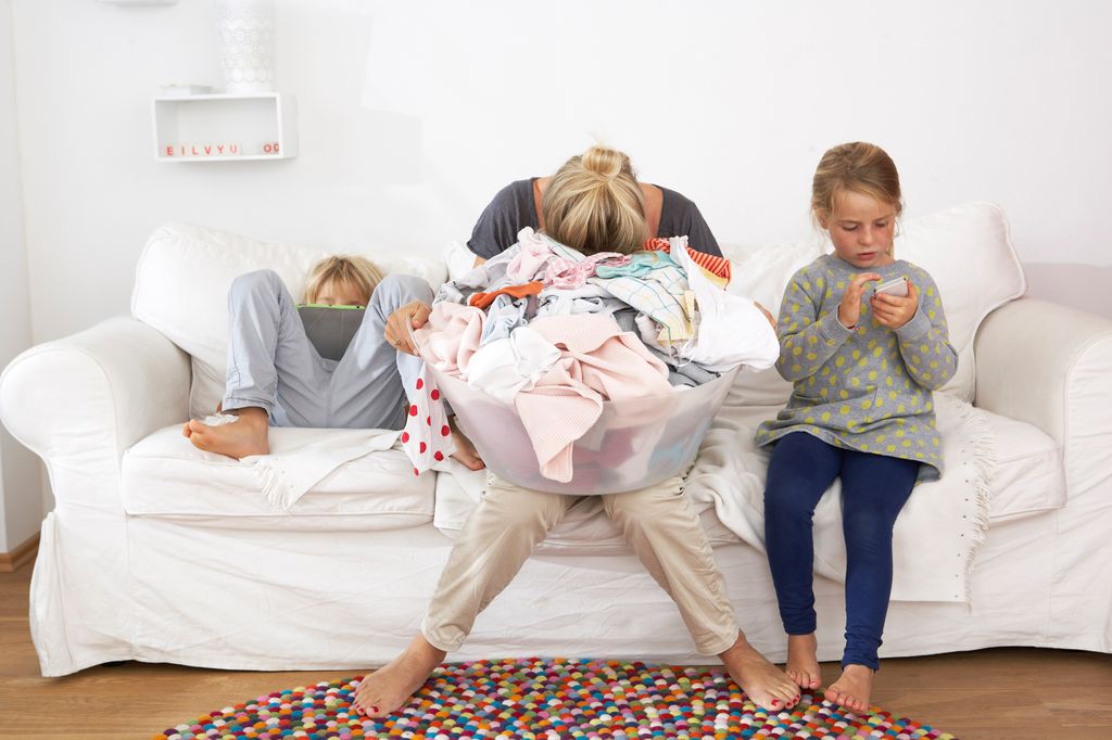 madre agotada, con la cesta de la ropa encima y sus hijos sentados en el sofá con el móvil