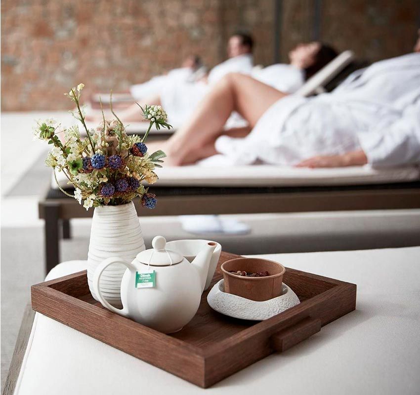 Un detalle de una bandeja con una infusión, al fondo clientes disfrutando del spa
