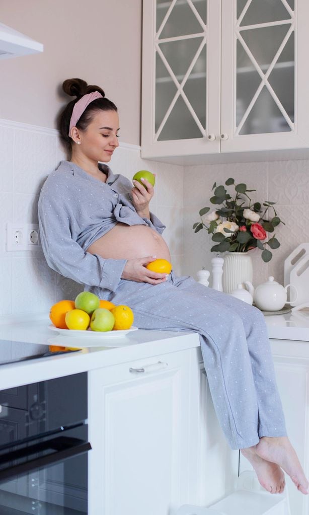 Embarazada sentada en la encimera de la cocina con una manzana en la mano