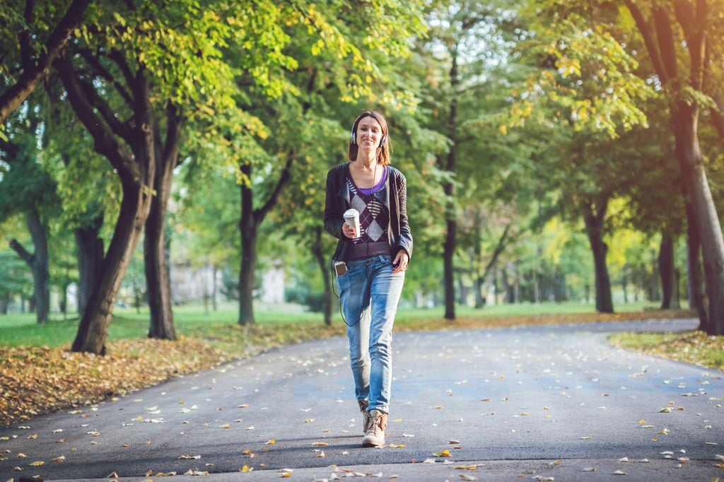 mujer caminando por un parque con los auriculares puestos y un café en la mano
