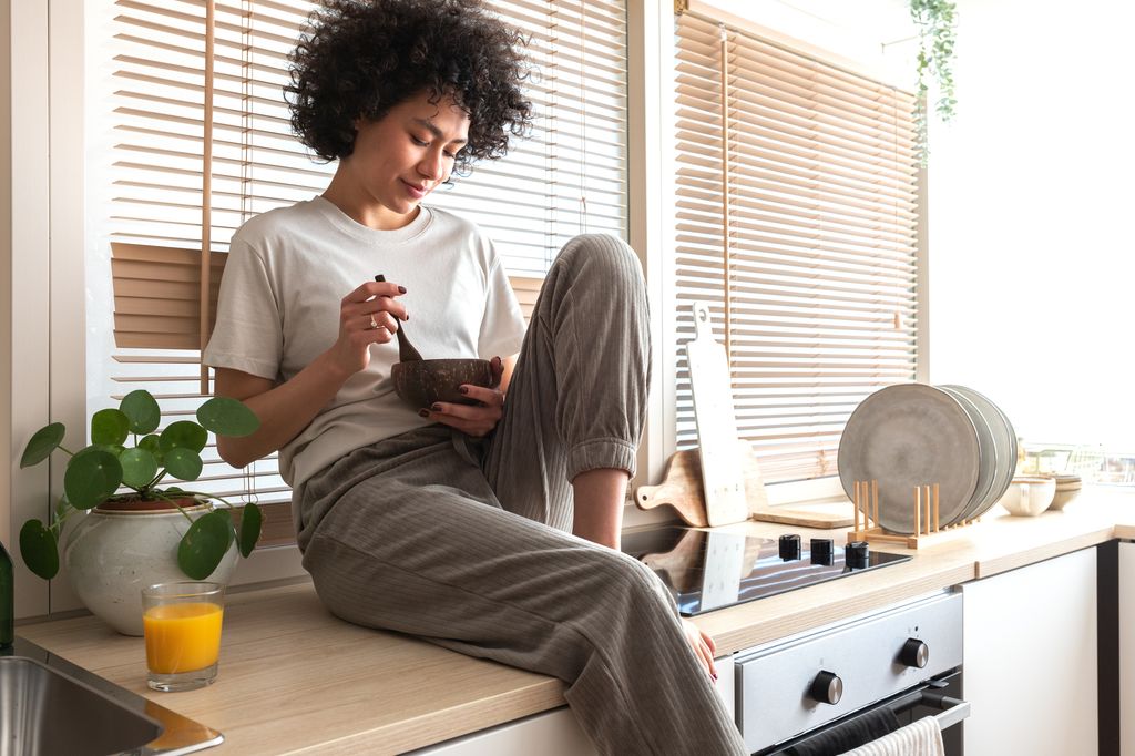 Mujer sentada en encimera de cocina con cuenco, vistiendo camiseta blanca y pantalones caqui.