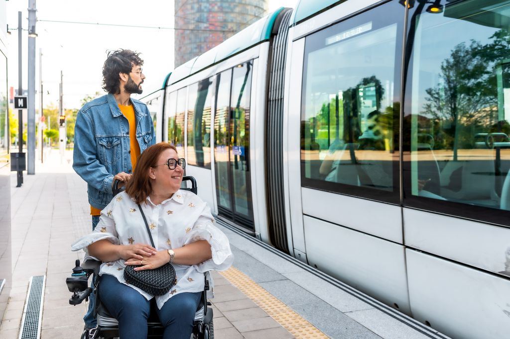 Un cuidador empuja a una mujer sonriente en silla de ruedas en una moderna estación de tranvía, ambos esperando abordar el transporte accesible.