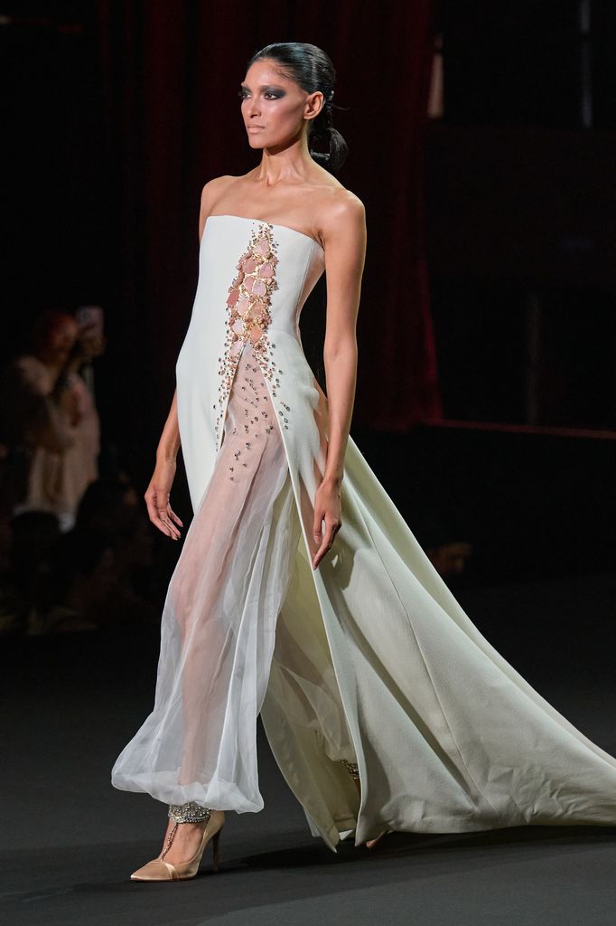 Modelo caminando en pasarela con vestido blanco y dorado, tacones dorados.