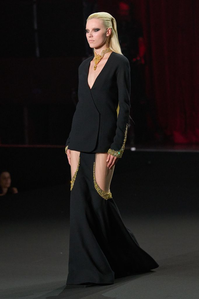Modelo rubia en traje negro con detalles dorados en pasarela.