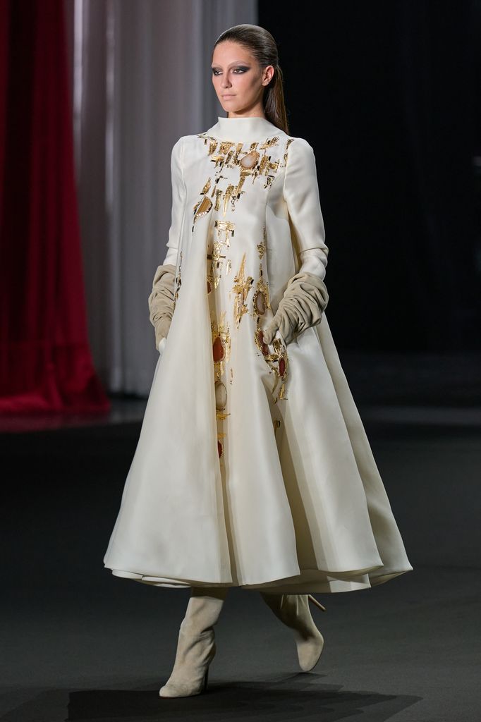 Modelo caminando en pasarela con vestido blanco y dorado, guantes y botas.