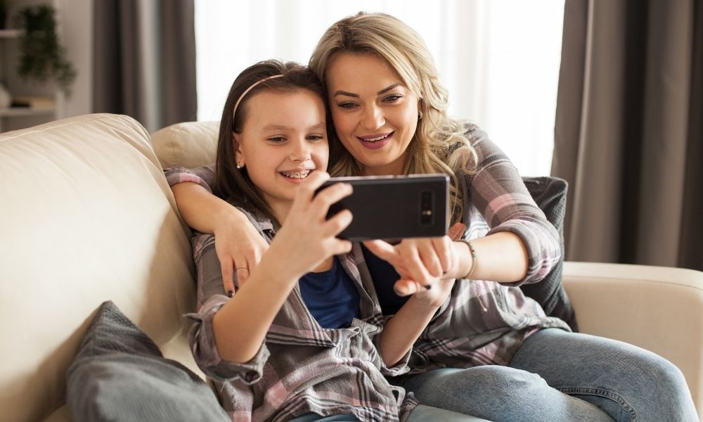 Madre e hija sonrientes revisando el teléfono móvil desde el sofá