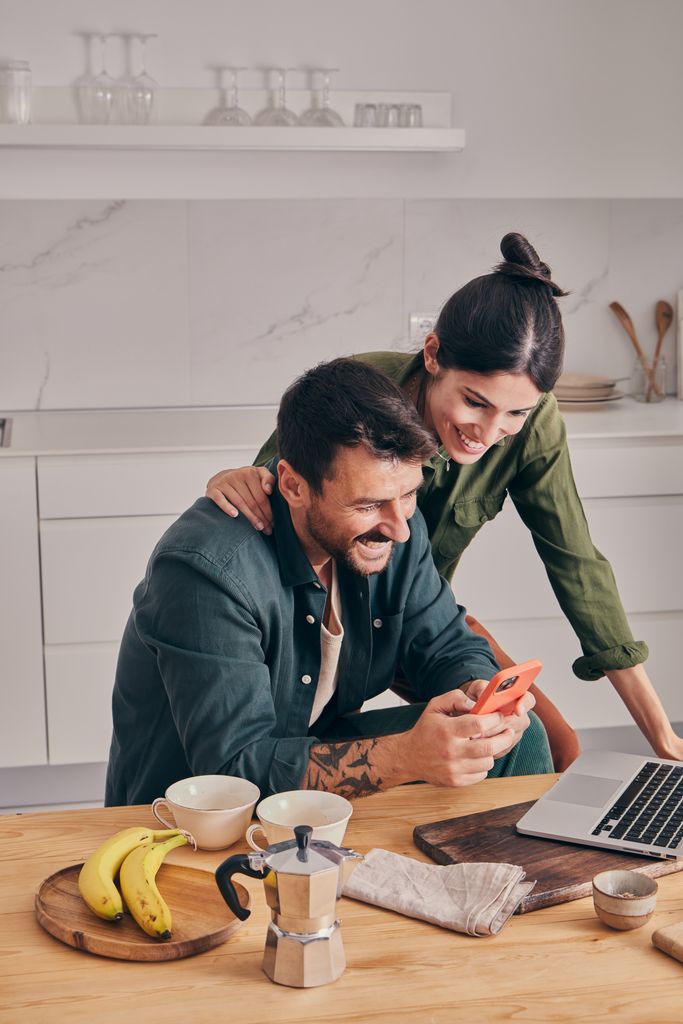 Una pareja con el movil y el ordenador sonriendo en la cocina