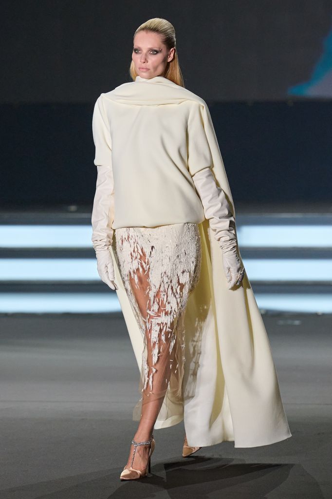 Anna Anka caminando en pasarela con traje blanco y guantes largos.