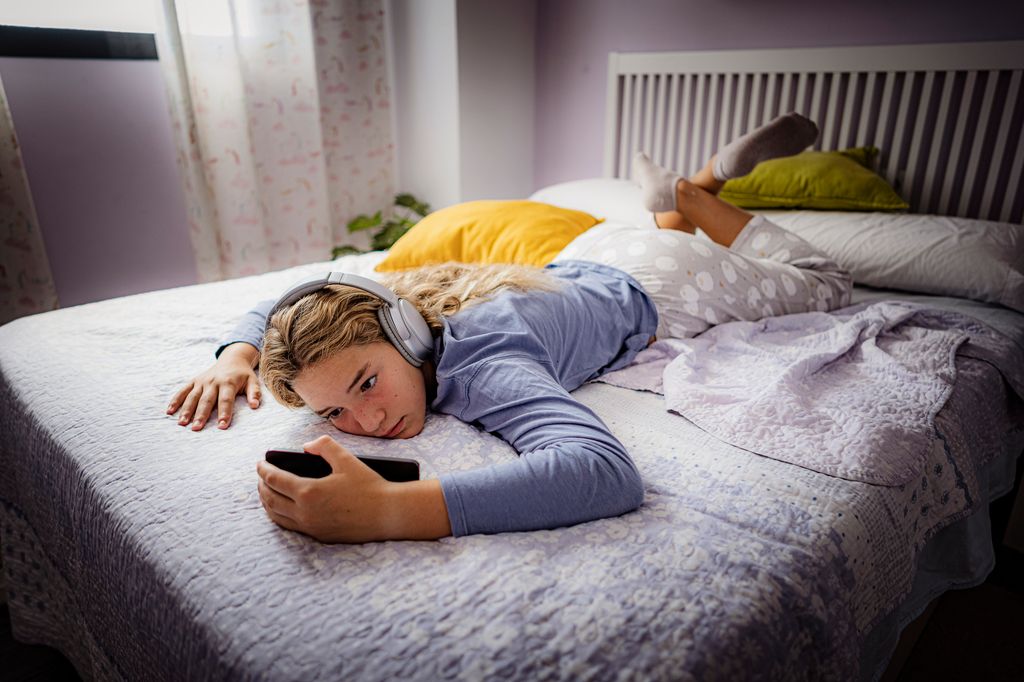 adolescente tumbada en la cama, escuchando música con los cascos puestos mientras mira al móvil