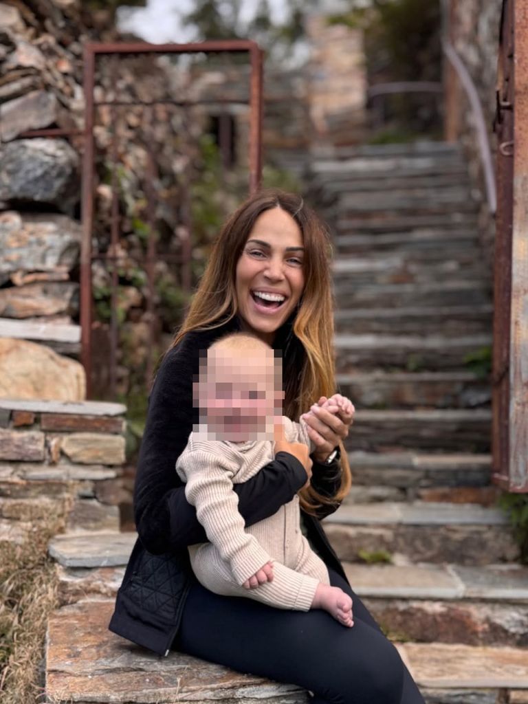 Mujer riendo con un bebé en sus brazos en un entorno rústico.