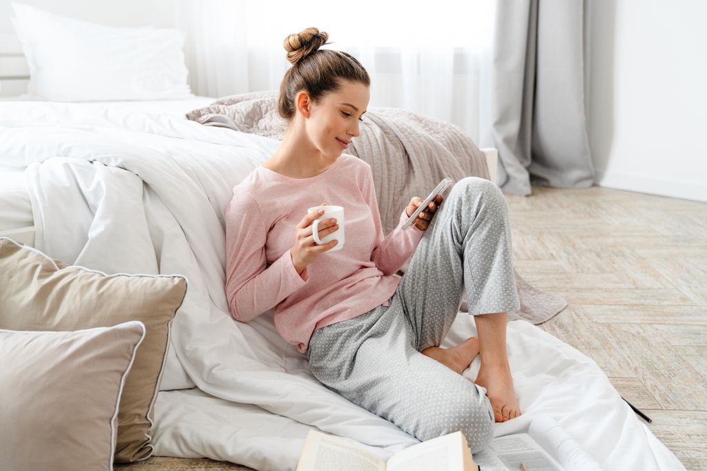 mujer joven mirando el móvil junto a la cama, tomando un café y con libros abiertos a sus pies