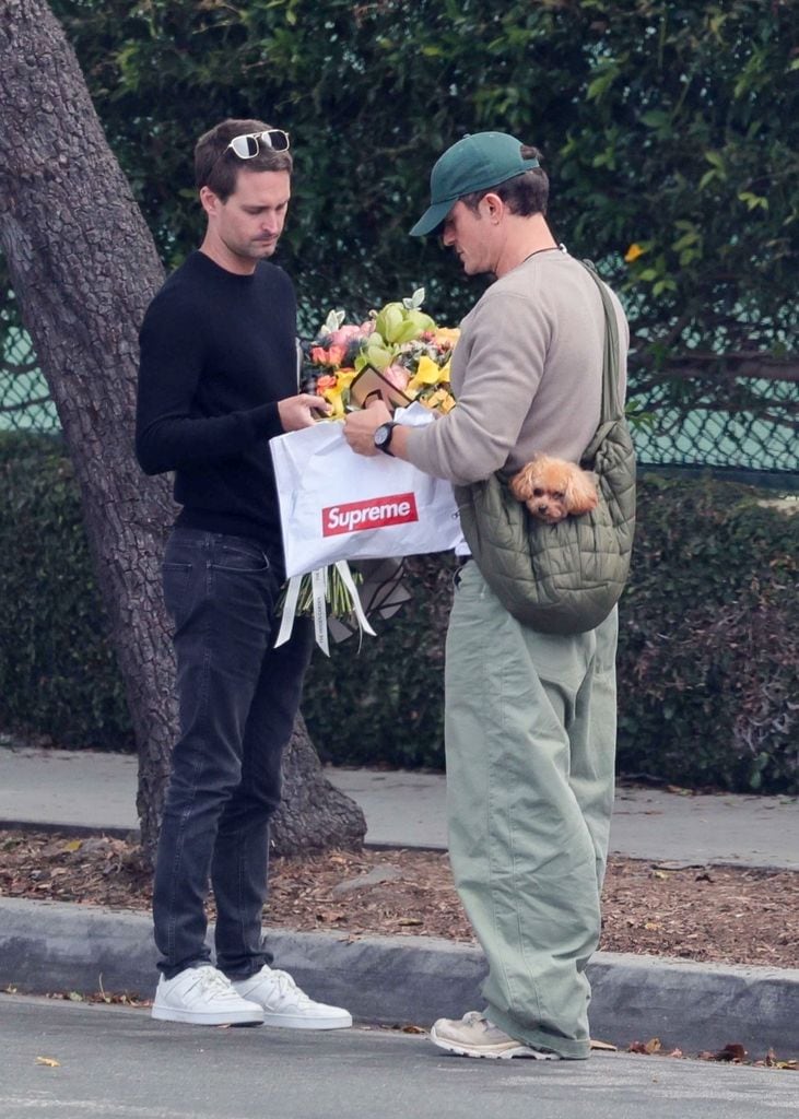 Evan Spiegel, vestido en tonos oscuros, con Orlando Bloom, en tonos claros y una gorra verde, conversan en la graduación de Flynn. 