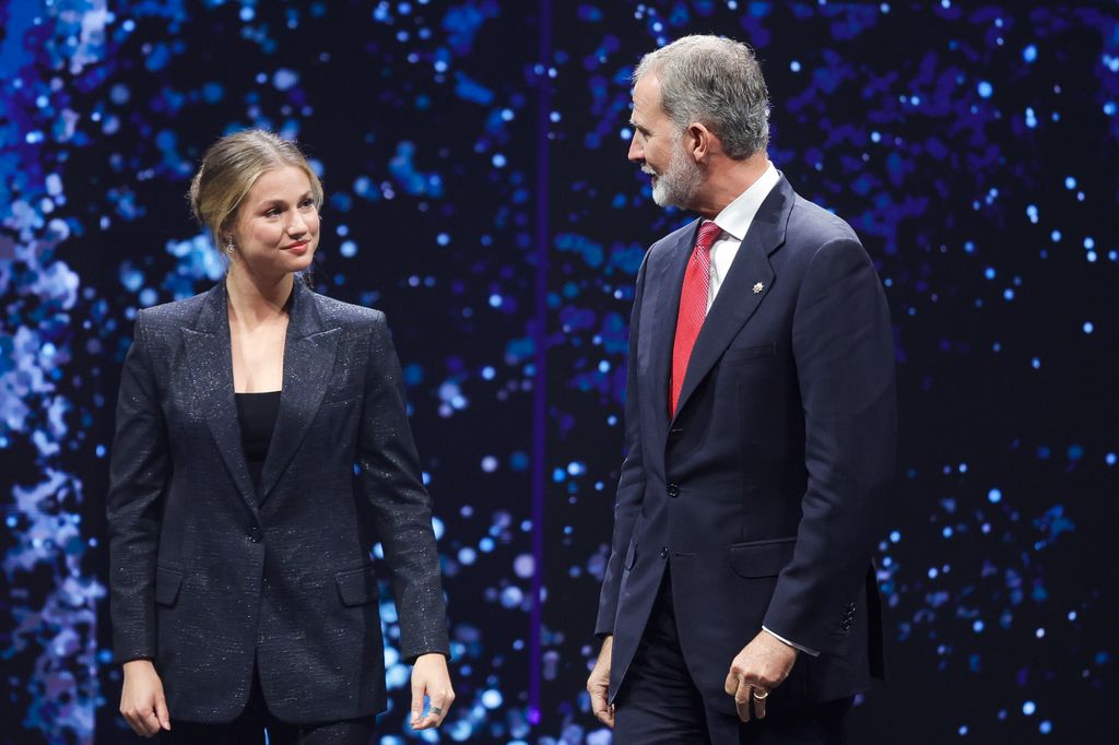 Mujer en traje negro y Felipe VI en traje azul con corbata roja conversan en escenario.