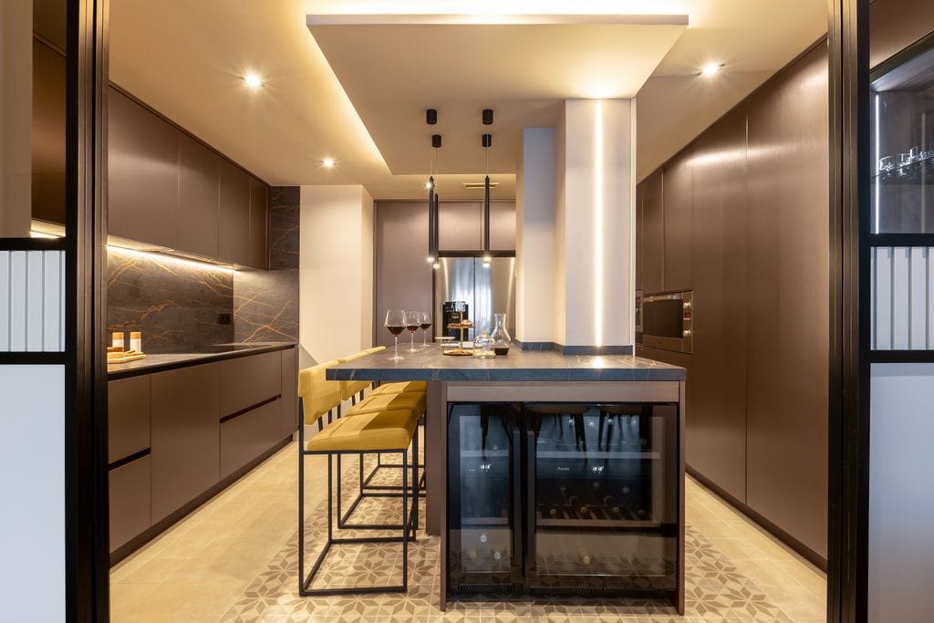 Cocina con isla central, vinoteca, taburetes tapizados amarillos, alfombra hidráulica, lámparas de techo negras y focos empotrados