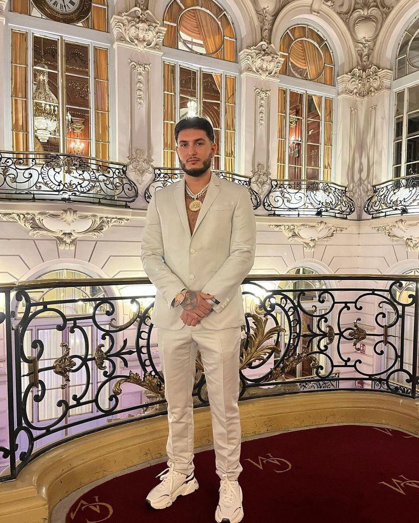 Hombre en traje blanco y zapatillas, posando en balcón lujoso con ornamentos dorados.