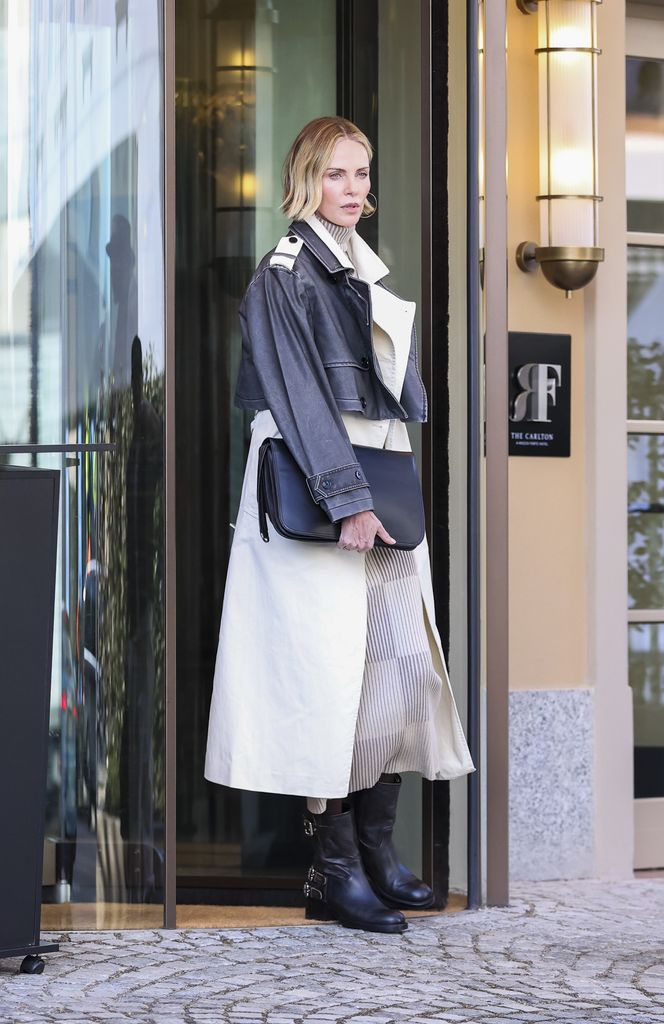 Lleva una gabardina larga color crema con una chaqueta corta de cuero oscuro superpuesta, combinada con un vestido midi tejido de cuello alto en tono neutro.