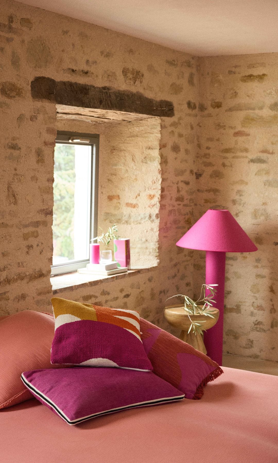 Dormitorio rústico con paredes de piedra, cama con ropa rosa salmón, cojines fucsias y lámparas pie fucsia