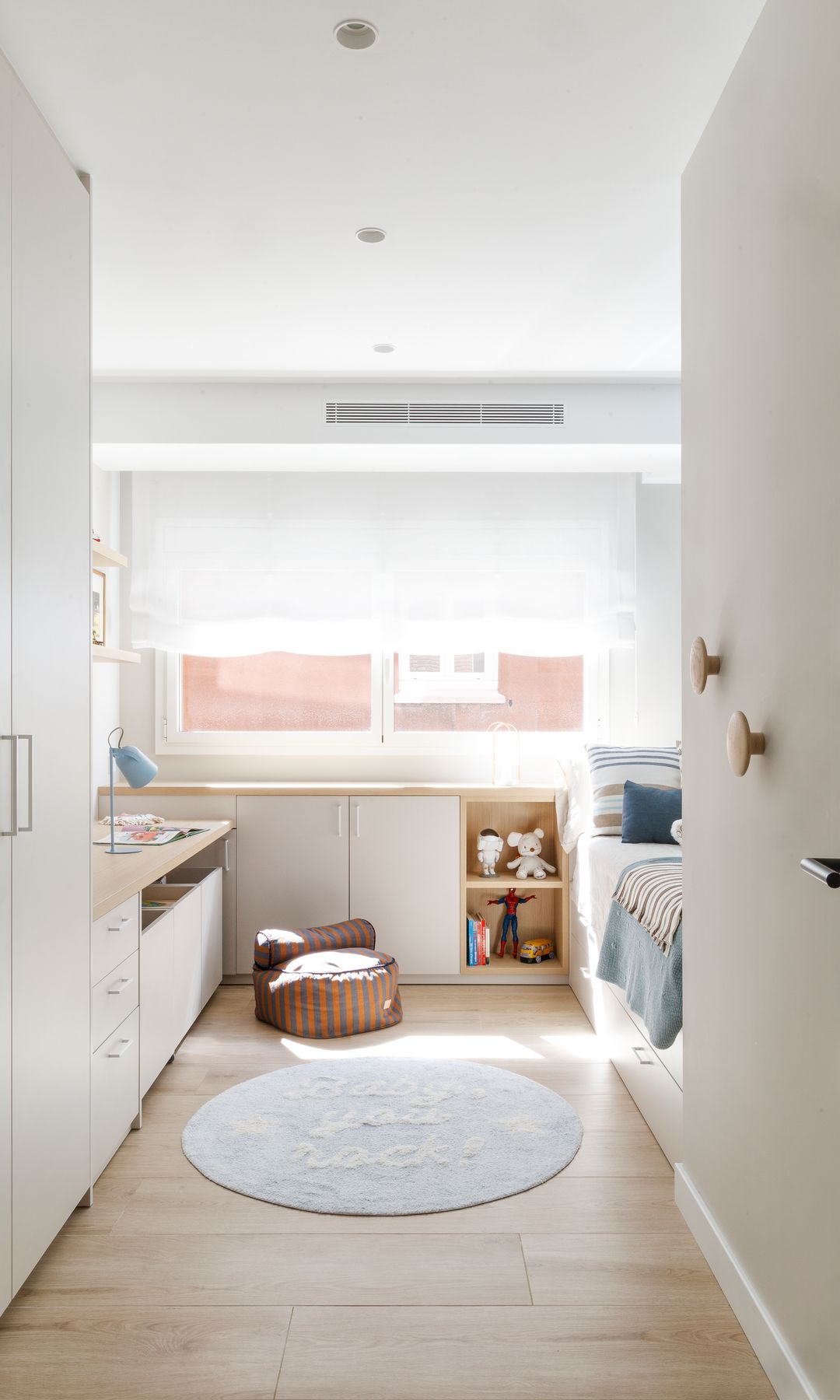 Habitación infantil con zona de escritorio, estantería baja bajo la ventana, alfombra redonda, puf, estores blancos, ropa de cama azul 