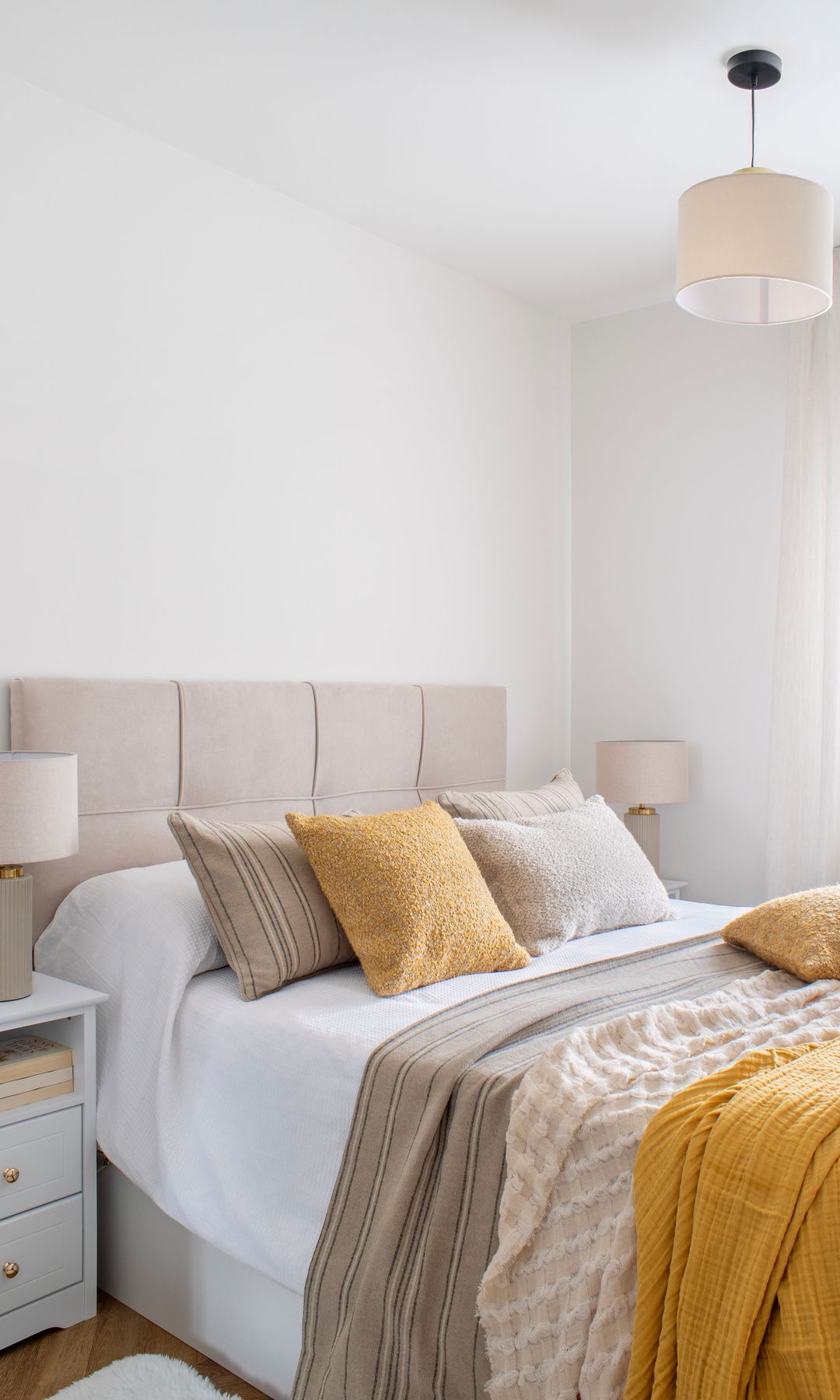 Dormitorio con cabecero tapizado en beige, ropa de cama beige y mostaza, cojines mostazas y beiges, lámpara de techo blanca, lámparas de sobremesa a juego sobre mesitas de noche