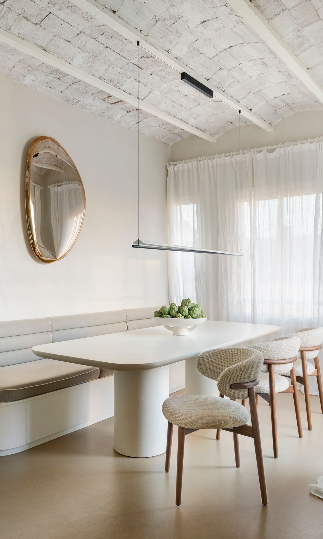 Comedor con mesa blanca, espejo en la pared, banco con respaldo tapizado, sillas de madera tapizadas, cortinas blancas